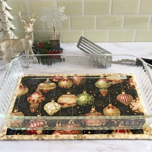 May include: A clear glass baking dish rests on a decorative Christmas-themed hot pad. The hot pad features a black background with gold, red, and green ornament designs. A second, smaller pad with a gold snowflake pattern is beneath.