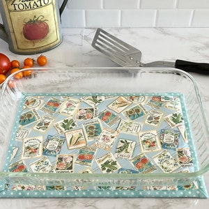 May include: A glass baking dish with a blue and white fabric hot pad underneath. The fabric has a vintage seed packet design with images of vegetables and flowers.