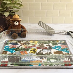 May include: A glass baking dish sits on a gray and white fabric hot pad with a colorful pattern of vintage-style national park signs. The signs feature images of bears, eagles, and other wildlife, along with text such as "Camping", "To Serpent's Trail", and "End of Trail". A stuffed ranger bear wearing a Smokey the Bear hat sits in the background.