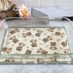 May include: A rectangular clear glass baking dish sits on a quilted fabric hot pad. The hot pad features a repeating pattern of brown highland cows and small flowers on a cream background with a sage green border and white daisy trim.