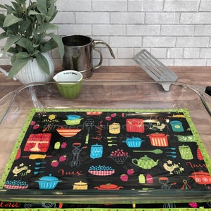 May include: A green and black kitchen themed fabric pot holder with a glass baking dish on top. The pot holder has a variety of kitchen items printed on it, including a teapot, a whisk, and a rolling pin.