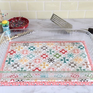 May include: A clear glass baking dish sitting on a quilted hot pad with a pink border. The hot pad has a patchwork design with various colors and patterns.