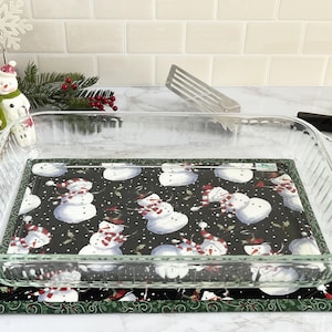 May include: A clear glass baking dish sits on a black and white snowman patterned kitchen mat with a green border.