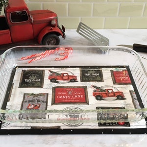 May include: A glass baking dish with a Christmas-themed fabric hot pad underneath. The hot pad features a red truck with a Christmas tree, the words "Candy Cane Factory", and other Christmas-themed designs.