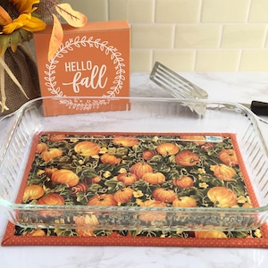 May include: A glass baking dish sits on a quilted hot pad with a pumpkin pattern. The hot pad has an orange border with white polka dots. The text "Hello Fall" is visible on a wooden sign in the background.