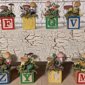 May include: Wooden alphabet blocks with decorative miniature mushroom and floral arrangements. Each block features a different letter in a contrasting color. The blocks are arranged on a distressed white surface, creating a whimsical display.