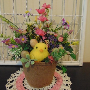 May include: A yellow chick figurine sits in a brown pot filled with pink, purple, and white flowers. The pot is on a white crocheted doily with a pink border.
