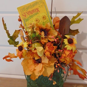 May include: A green metal container holds an arrangement of orange and yellow artificial flowers, green leaves, a small metal shovel, and a yellow seed packet labeled NK Lawn & Garden Co. Sunflower Giant Greystripe with a net weight of 6.5g.