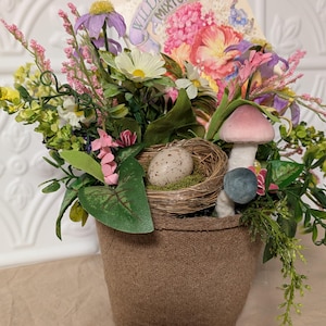 May include: A small burlap pot filled with a variety of pink, white, and green flowers. A bird's nest with a speckled egg sits in the center of the arrangement. A vintage seed packet for wildflower mix is visible in the background.