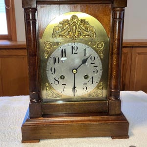 May include: A wooden antique clock with a gold-colored face and decorative accents. The clock has a rectangular shape with a curved top and two columns on either side of the face. The clock face displays numbers and hands. Includes a key and pendulum.