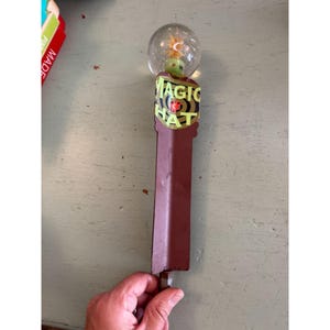 May include: A brown and yellow Magic Hat tap handle with a clear globe top. The handle has the words "MAGIC HAT" in yellow and black. The globe contains a small green figure and a yellow starburst.