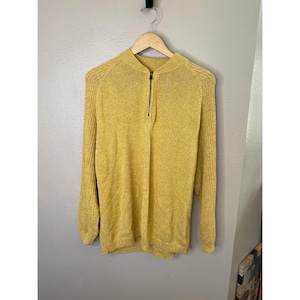 May include: Mustard yellow sweater with a half-zip closure. The long-sleeved sweater is hanging on a wooden hanger against a neutral wall. The sweater has a ribbed texture and a high neckline.