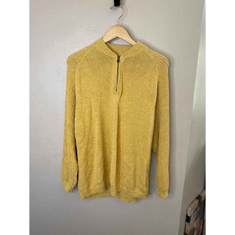 May include: Mustard yellow sweater with a half-zip closure. The long-sleeved sweater is hanging on a wooden hanger against a neutral wall. The sweater has a ribbed texture and a high neckline.