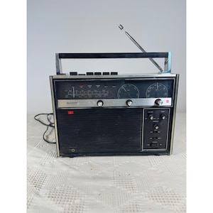 Può includere: Una radio portatile vintage Sharp solid state con corpo rettangolare nero, pannello di controllo argentato con manopole di sintonia, griglia dell'altoparlante nera, maniglia superiore per il trasporto e un'antenna metallica estensibile.