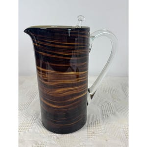 May include: A cylindrical glass pitcher featuring a dark brown and amber horizontal swirl pattern. The vessel includes a clear glass handle attached to the side and a small clear glass stirring rod resting inside the top opening of the pitcher.