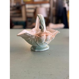 Puede incluir: Una pequeña cesta de cerámica con asa, en tonos verde claro y melocotón. La cesta tiene un diseño de hojas y se apoya sobre una base de pedestal. El asa es arqueada y el interior es blanco. Altura aproximada 15 cm.