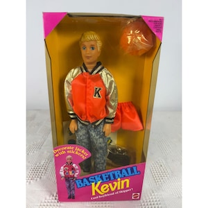 May include: A Basketball Kevin doll in original packaging, featuring a blonde male doll wearing an orange and gold varsity jacket with a letter K, and grey patterned pants. The box includes an orange basketball and additional orange shorts.
