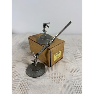 May include: A pair of silver-toned metal soldering stands with circular weighted bases and adjustable articulated arms featuring wing nuts. The stands are positioned in front of a rectangular cardboard box with a yellow label containing text.