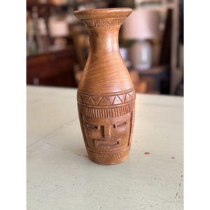 May include: A light brown wooden vase with carved tribal face and geometric patterns. The vase has a wide base, a cylindrical body, and a flared top. The face is carved into the front of the vase, with a rectangular mouth and eyes.