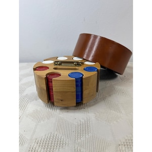 May include: A round wooden poker chip carousel with a brass handle, holding stacks of red, blue, and white plastic chips. A separate, smooth brown cylindrical lid sits tilted to the side, revealing the organized storage compartments.