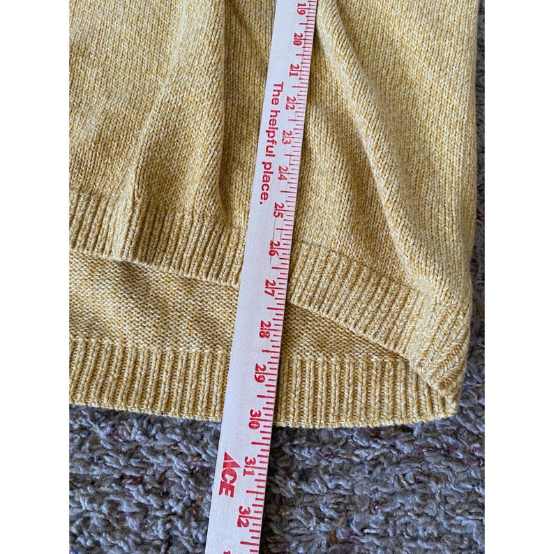 May include: Close-up of a mustard yellow sweater with a ribbed hem. A measuring tape is placed vertically across the sweater, showing measurements from 19 to 32 inches. The text "The helpful place" is printed on the tape.