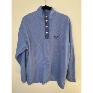 May include: A blue long-sleeve pullover shirt with a fine vertical pinstripe pattern, a mock neck collar, and a four-button placket. The shirt features the text FILA SPORT embroidered in dark blue on the upper right chest area.