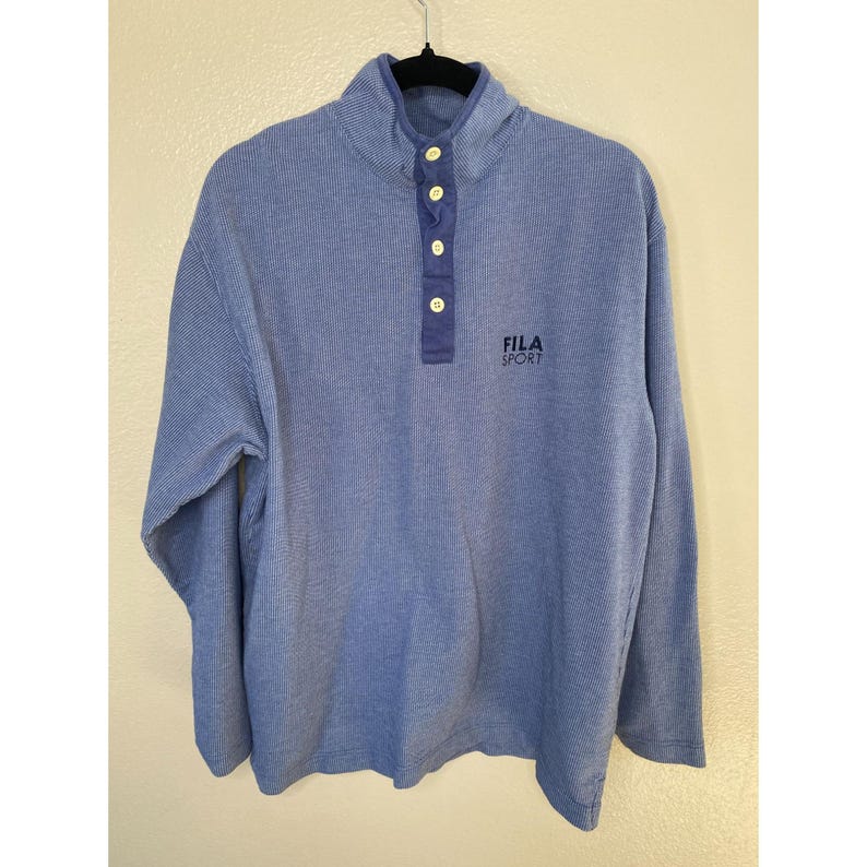 May include: A blue long-sleeve pullover shirt with a fine vertical pinstripe pattern, a mock neck collar, and a four-button placket. The shirt features the text FILA SPORT embroidered in dark blue on the upper right chest area.