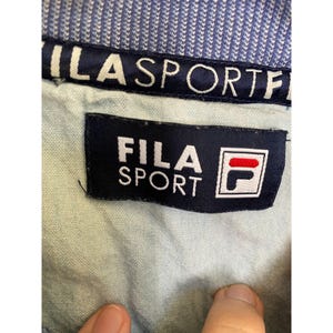 May include: A light blue garment featuring a dark blue interior collar band with the white text FILA SPORT. A rectangular dark blue fabric label is sewn onto the light blue fabric, displaying the white text FILA SPORT and the red and blue F logo.