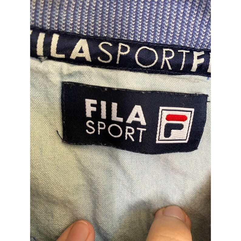 May include: A light blue garment featuring a dark blue interior collar band with the white text FILA SPORT. A rectangular dark blue fabric label is sewn onto the light blue fabric, displaying the white text FILA SPORT and the red and blue F logo.