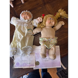 Könnte beinhalten: Zwei Cabbage Patch Kids Puppen. Eine Puppe trägt ein gelbes Kleid mit Spitzenbesatz und eine Haube. Die andere Puppe trägt eine Windel mit Blumenmuster. Beide Puppen liegen auf Adoptionsurkunden.