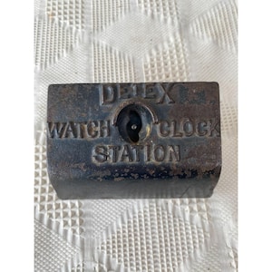 Pode incluir: Um objeto metálico retangular e desgastado com a inscrição em relevo "DETEX WATCH CLOCK STATION". Apresenta um buraco de fechadura e provavelmente é um componente de uma antiga estação de relógios. Aspeto escuro e envelhecido.