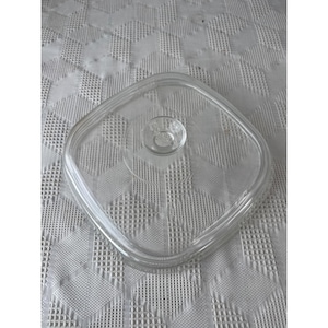 Könnte beinhalten: Ein klarer, quadratischer Glasdeckel mit abgerundeten Ecken und einem kleinen, kreisförmigen Griff in der Mitte. Der Deckel ist für eine quadratische Schüssel konzipiert. Das Glas ist transparent.