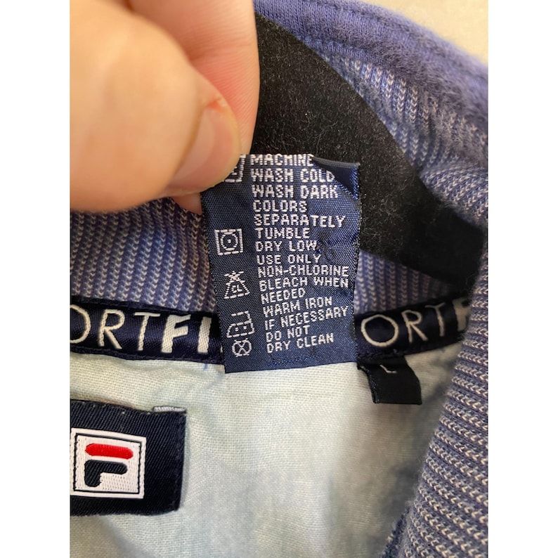 May include: A blue Fila garment featuring a dark blue interior care label with white text that reads MACHINE WASH COLD, WASH DARK COLORS SEPARATELY, TUMBLE DRY LOW, USE ONLY NON-CHLORINE BLEACH WHEN NEEDED, WARM IRON IF NECESSARY, DO NOT DRY CLEAN.