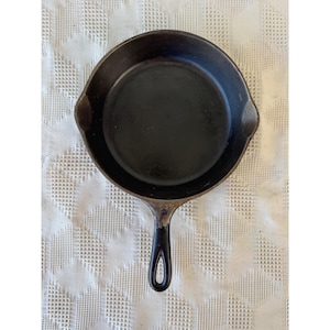 May include: A round black cast iron skillet with a smooth interior surface and a single long handle featuring a teardrop-shaped hole at the end. The pan includes two small pouring spouts on opposite sides of the rim for liquid drainage.