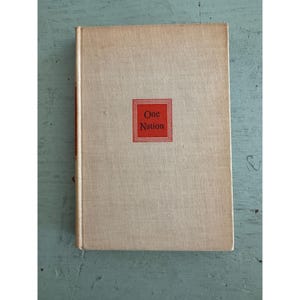 May include: A hardcover book with a light beige fabric cover. Centered on the front cover is a red square frame containing the text One Nation in black lettering. The book has a vertical red stripe along the spine edge of the front cover.