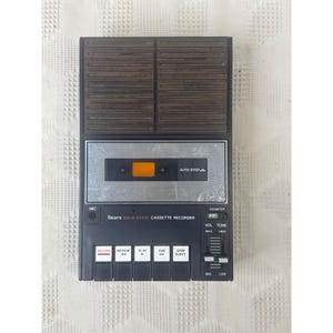 May include: A black and brown Sears Solid State cassette recorder featuring a top speaker grille, a central cassette compartment with an orange window, and a control panel with buttons for record, review, play, cue, and stop/eject functions.