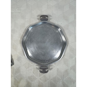 Puede incluir: Una bandeja de servicio hexagonal de metal plateado con un acabado liso y cepillado en la superficie central. La bandeja cuenta con dos asas opuestas con un acabado oscuro, texturizado y martillado, y un detalle recortado en el centro.