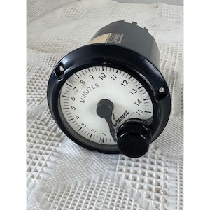 Puede incluir: Un temporizador analógico en blanco y negro con esfera circular, que presenta una escala de 1 a 15 minutos. El dispositivo tiene una carcasa exterior negra, una esfera blanca con números negros, una aguja central y un mando de control negro con la inscripción Cramer U.S.A.