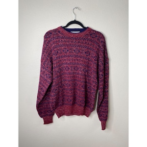 May include: A maroon and navy blue patterned sweater hangs from a black hanger. The sweater has a crew neck with a navy blue collar. The knitwear features a textured pattern in shades of maroon and navy blue. The sleeves are long and the cuffs are ribbed.
