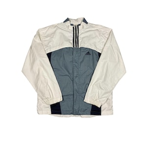 May include: A white, gray, and navy blue windbreaker jacket featuring a high collar, long sleeves with elastic cuffs, and a front zipper closure. The Adidas logo is embroidered in navy blue on the upper chest, with three vertical stripes.