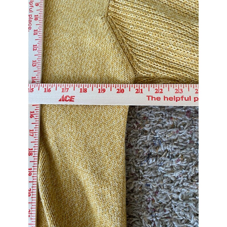 May include: Close-up of a mustard yellow knit sweater. A ruler is placed across the sweater, measuring from 16 to 23 inches. The text "ACE" and "The helpful place" are visible on the ruler.