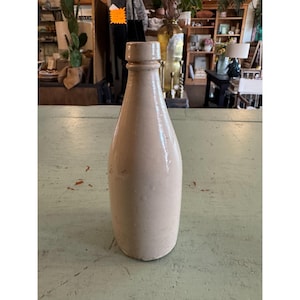 May include: A vintage, off-white stoneware bottle with a narrow neck and rounded body. The bottle has a slightly textured surface and a light brown hue. It is likely an antique piece, suitable for display or as a decorative element.