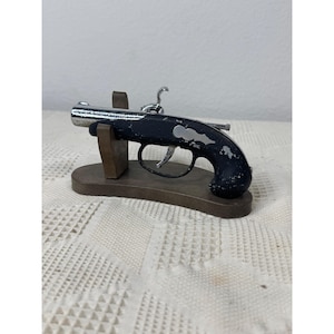 以下が含まれることがあります： 黒いハンドルに銀色のアクセントと金属製の銃身を備えた、フリントロック式ピストルのミニチュアレプリカ。ピストルは湾曲した茶色の木製スタンドに取り付けられており、詳細なトリガーメカニズムと装飾的なサイドプレートが特徴です。
