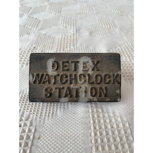 Pode incluir: Uma placa retangular de metal desgastado com o texto "DETEX WATCHCLOCK STATION" em relevo na sua superfície. A placa é de cor cinza escuro com algumas áreas de cinza mais claro, e repousa sobre uma superfície branca texturizada.