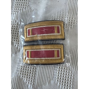 May include: Two rectangular shoulder boards featuring a central red fabric panel bordered by white and gold metallic embroidery. Each piece has two vertical gold bars on the red section and is enclosed in a clear plastic protective sleeve.
