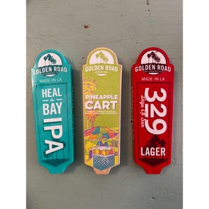 May include: Three wooden beer tap handles from Golden Road Brewing. The handles are teal, green, and red, with the brewery's logo and various beer names: "Heal the Bay IPA", "Pineapple Cart", and "329 Lager".