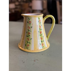 May include: A yellow ceramic pitcher with vertical stripes and a heart and vine design. The pitcher has a curved handle and a spout for pouring. The design features red hearts and green vines. The pitcher is likely used for serving beverages.