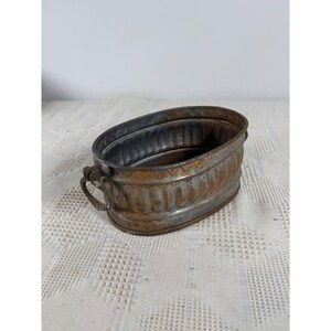 May include: An oval-shaped metal basin with a weathered copper and gray finish. The exterior features a vertical ribbed pattern and a decorative band near the rim. Two small, twisted metal handles are attached to the sides of the container.
