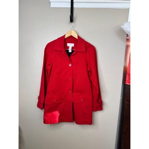 May include: A bright red button-down jacket with a classic collar, long sleeves with cuff tabs, and two large front flap pockets. The garment features a visible white brand label at the inner neckline and is displayed on a wooden hanger.