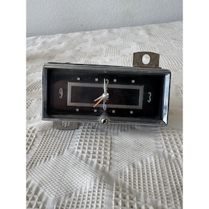May include: A rectangular analog dashboard clock with a black face, silver trim, and white numerals at the 3, 6, and 9 positions. The clock features white hour and minute hands, a thin orange second hand, and a small adjustment knob at the bottom.